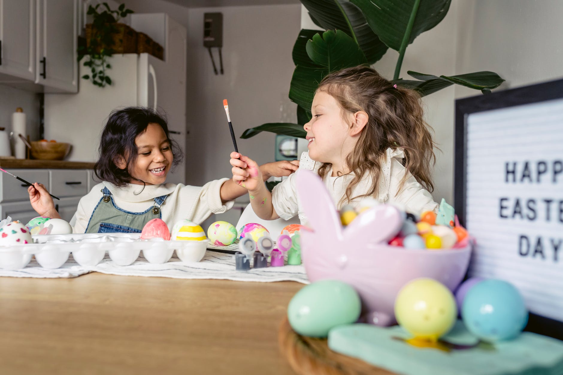 happy multiethnic girls painting eggs on table in kitchen
